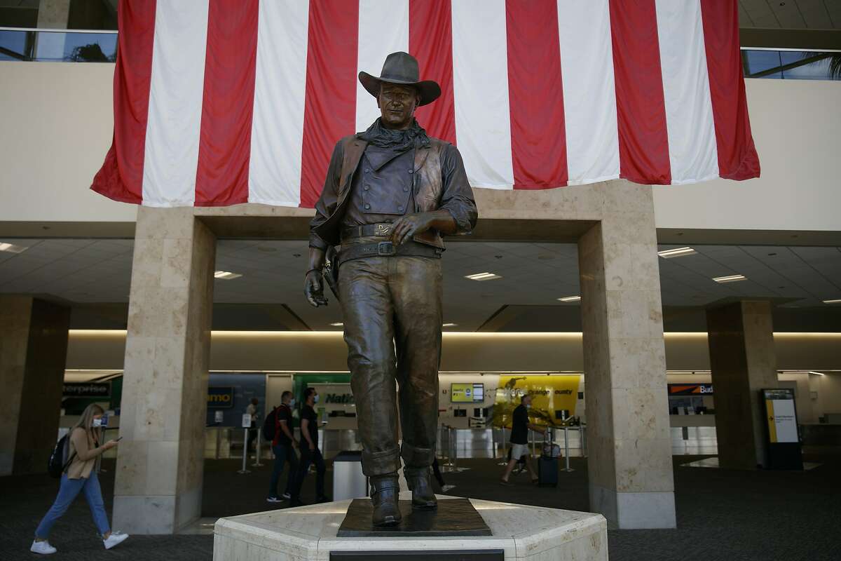 Travelers walk past a John Wayne statue at John Wayne Airport in Santa Ana, Calif, Monday, June 29, 2020. In the latest move to change place names in light of U.S. racial history, leaders of Orange County's Democratic Party are pushing to drop film legend Wayne's name, statue and other likenesses from the county's airport because of his racist and bigoted comments. (AP Photo/Jae C. Hong)