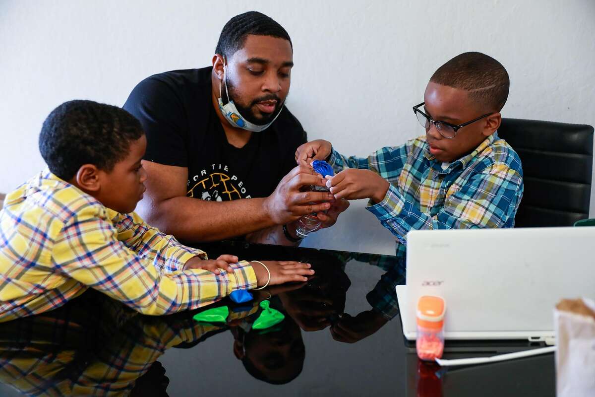 James Thomas helps his son Jamal Lee Jr., 10, re-create a lung with a balloon and plastic bottle during an online science class at their San Leandro home as his other son, Javaughn Thomas, 7, watches.