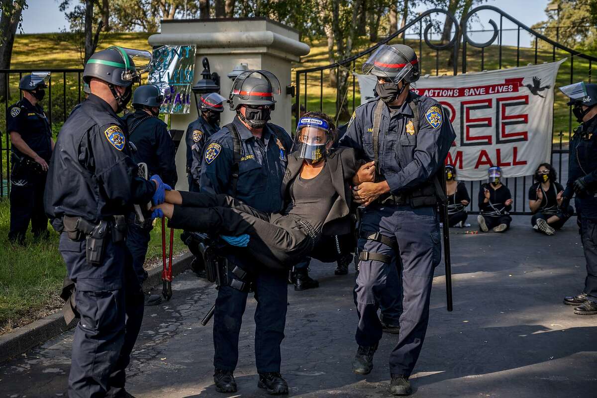 A woman protester is carried away by California Highway Patrol officers outside the gate of California Gov. Gavin Newsom's home in Fair Oaks, Calif., on Monday, July 27, 2020. Demonstrators chained themselves to a fence outside the governor's home, calling for mass inmate releases and an end to immigration transfers because of the coronavirus pandemic, as deaths mounted at a San Francisco Bay Area prison. The CHP cut the chains linking protesters to the bars of the gate at the front of the residence in suburban Sacramento after about two hours, but could not immediately say how many had been arrested. (Daniel Kim/The Sacramento Bee via AP)