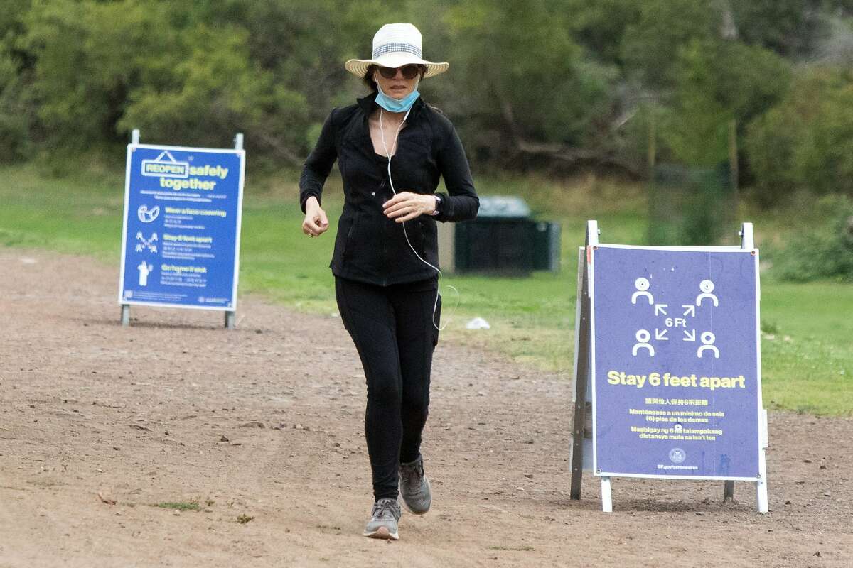 A woman jogging in Golden Gate Park runs past signs reminding people at a social distance and wearing masks in San Francisco on July 28, 2020.