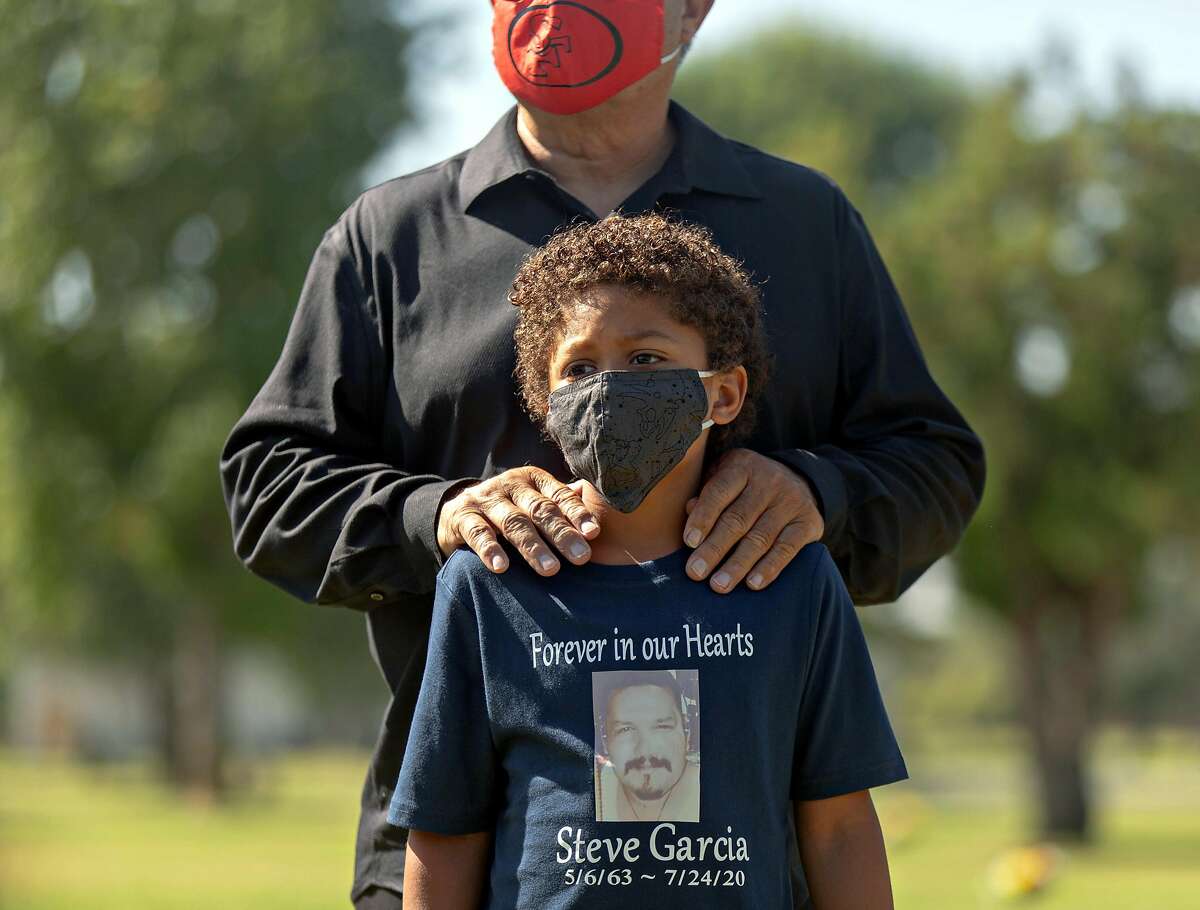 Raymond Price, front, stands with his grandfather Jesse Vargas during the services for Steven Garcias, who died of Covid-19 last month at Avenal State Prison, at San Fernando Mission Cemetery in Los Angeles, Calif. on Friday, August 7, 2020. Several relatives of men who died of Covid-19 in California prisons have gotten a rude shock during the mourning process: The state is making them pay thousands of dollars for the burial or cremation of their loved ones.