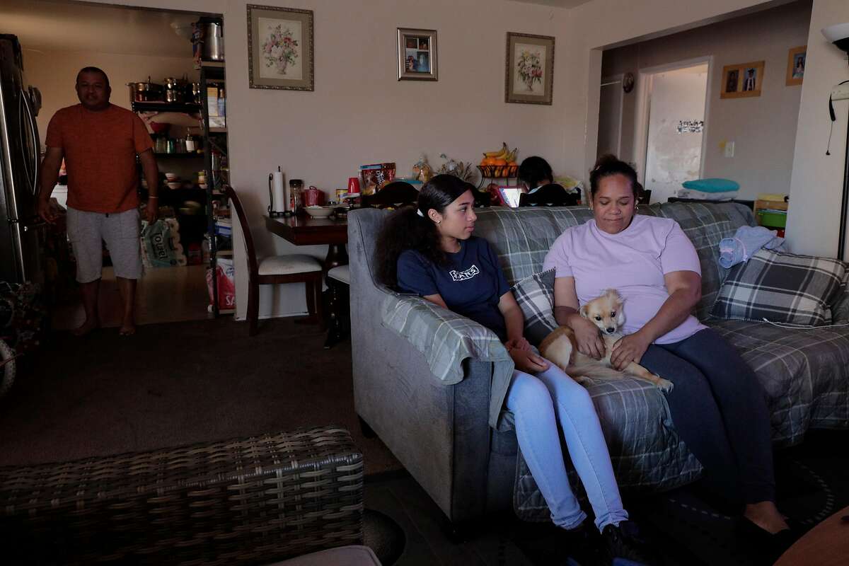 Alfredo Romero, left, walks from the kitchen to the living room where his wife, Elena Sabay, right, and daughter, Johmara, 14, are seated as the Romero family spends time at home in Oakland, Calif., on Monday, July 13, 2020. Elena Sabay recently lost her job as hotel housekeeper because of business shutdowns and her husband’s, Alfredo Romero, earnings are the only thing keeping the family afloat during the pandemic.