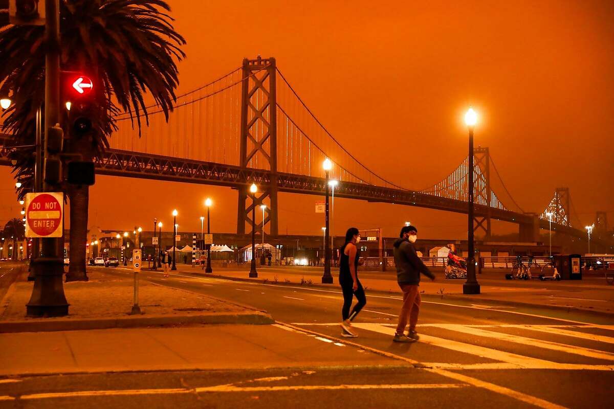 People pass by the Bay Bridge as dark orange skies hang over downtown San Francisco because of multiple wildfires burning across California and Oregon on Sept. 9.