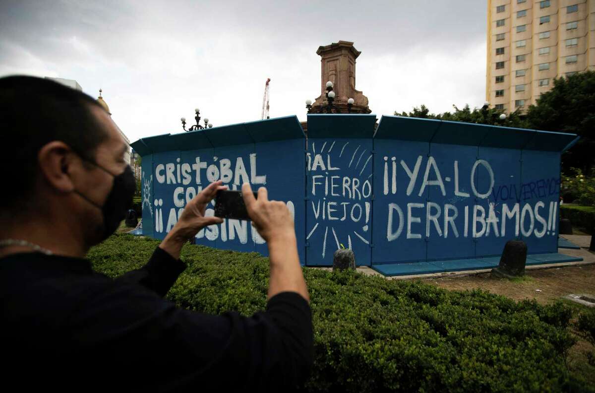 Un peatón toma una foto de un grafiti con la leyenda “Cristóbal Colón asesino, ya lo derribamos”, en una barrera temporal colocada para proteger el perímetro de la estatua de Cristóbal Colón, que fue retirada por las autoridades en el Paseo de la Reforma de la Ciudad de México, el lunes 12 de octubre de 2020.