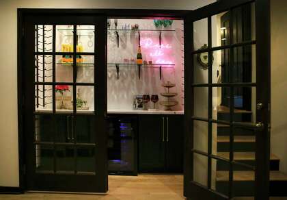 A cocktail nook, located where the kitchen used to be. Fronted by two glass doors that originally served as back doors to the house, the closet looks like a storefront display of wine-related wares with a wine cooler, bottle hangers and a pink neon sign reading, “Rosé all day.”