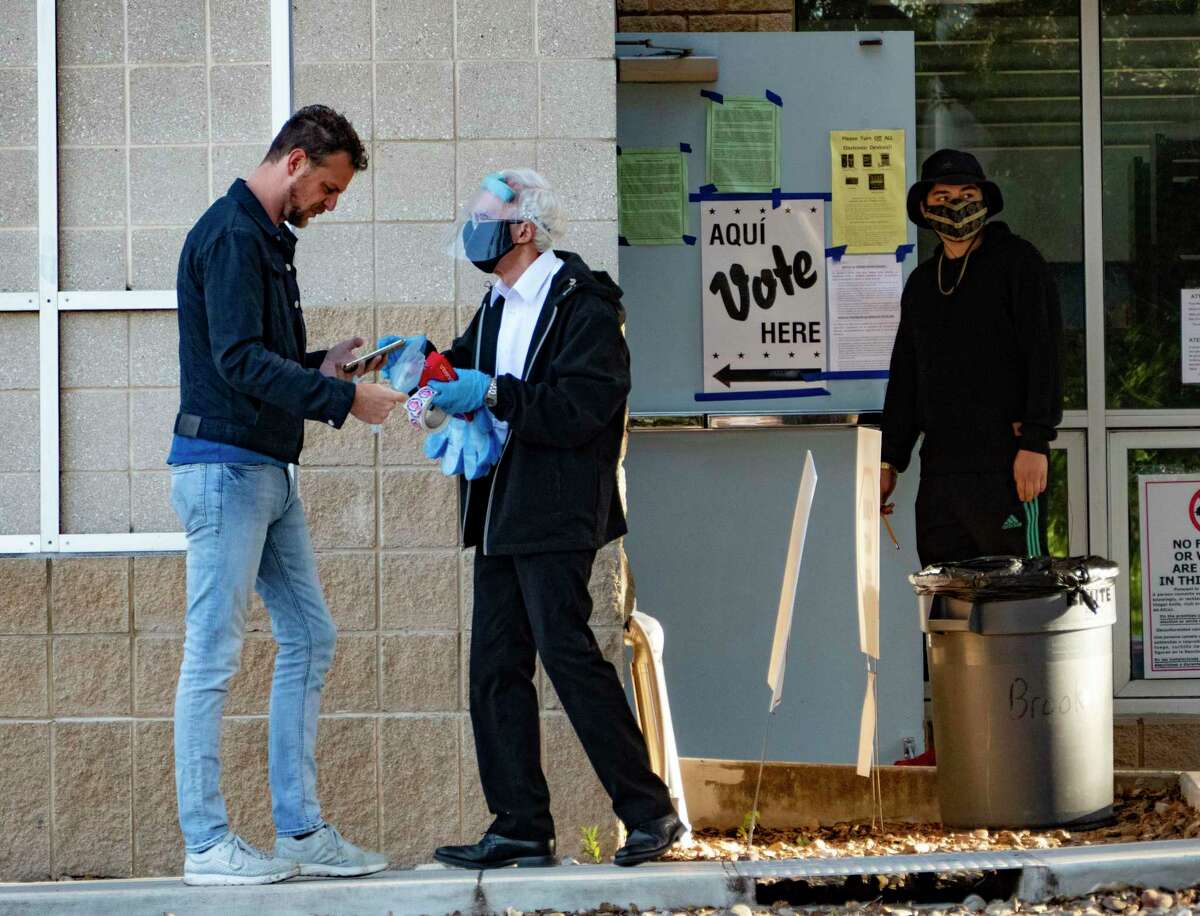 An election official, clad in protection against the spread of the coronavirus, offers gloves to a voter at the Brook Hollow Branch Library early on Election Day.