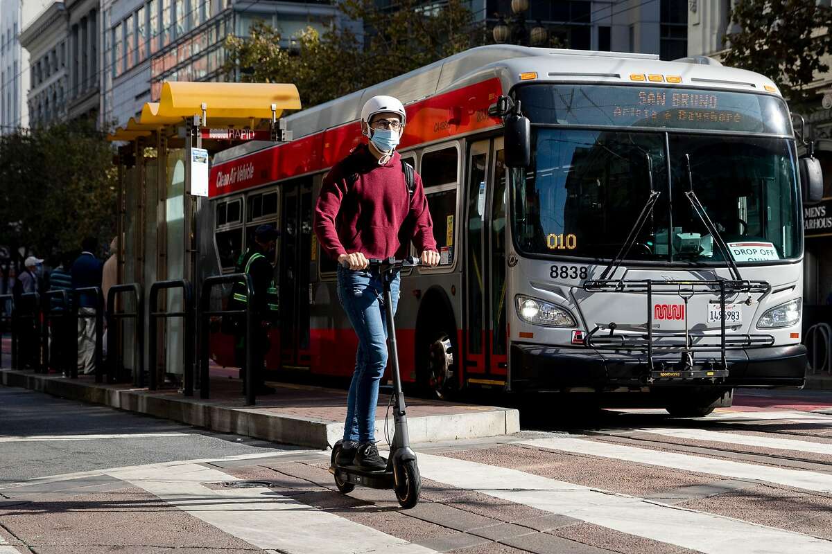 A person on a scooter move past a bus near 4th and Market street in San Francisco, Calif. Friday, October 30, 2020. The Department of Public Works and the San Francisco Municipal Transportation Authority has been quietly telling stakeholders the past couple weeks that due to budget cutbacks, bicycles will now share a lane along with taxis, paratransit, and commercial vehicles, and Muni busses will use only the center lane instead of curbside stops. The city will save $40 million, but it will still cost $121 million and an additional $7 million for redesign. The plane came to a head at a meeting Tuesday, with city supervisors and bike and pedestrian advocates protesting the sudden change.