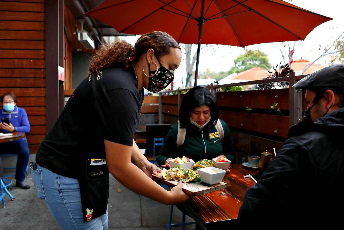 Server Briselle Lopez delivers food to diners at 5 Tacos and Beers on Solano Ave. on Saturday, December 5, 2020, in Albany, Calif. The Bay Area is expected to go under lockdown and sheltering in place on Sunday. Tipped employees, like servers and bartenders, can be paid as low as $2.13 per hour in some states. The Extra Spicy podcast explores the link between America’s tipping culture and the minimum wage debate, and why the fight for fair wages is not just a “restaurant issue.”