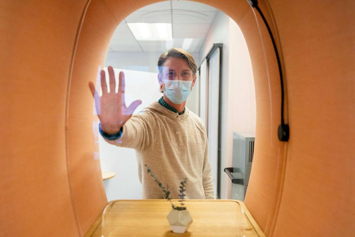 Brandon Cook places his hand on a plexiglass barrier at a standing desk at Workplace 2030 in San Francisco.