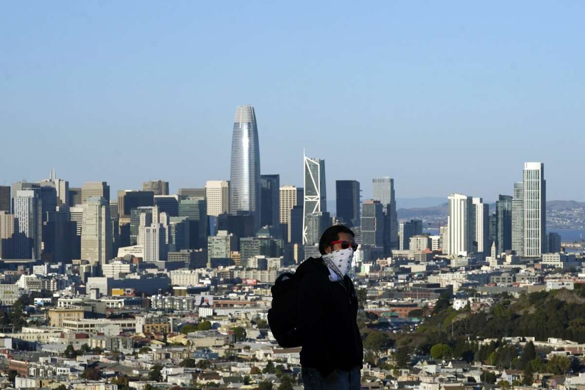 A person wearing a protective mask walks in front of the skyline on Bernal Heights Hill amid the coronavirus pandemic in San Francisco, Monday, Dec. 7, 2020.