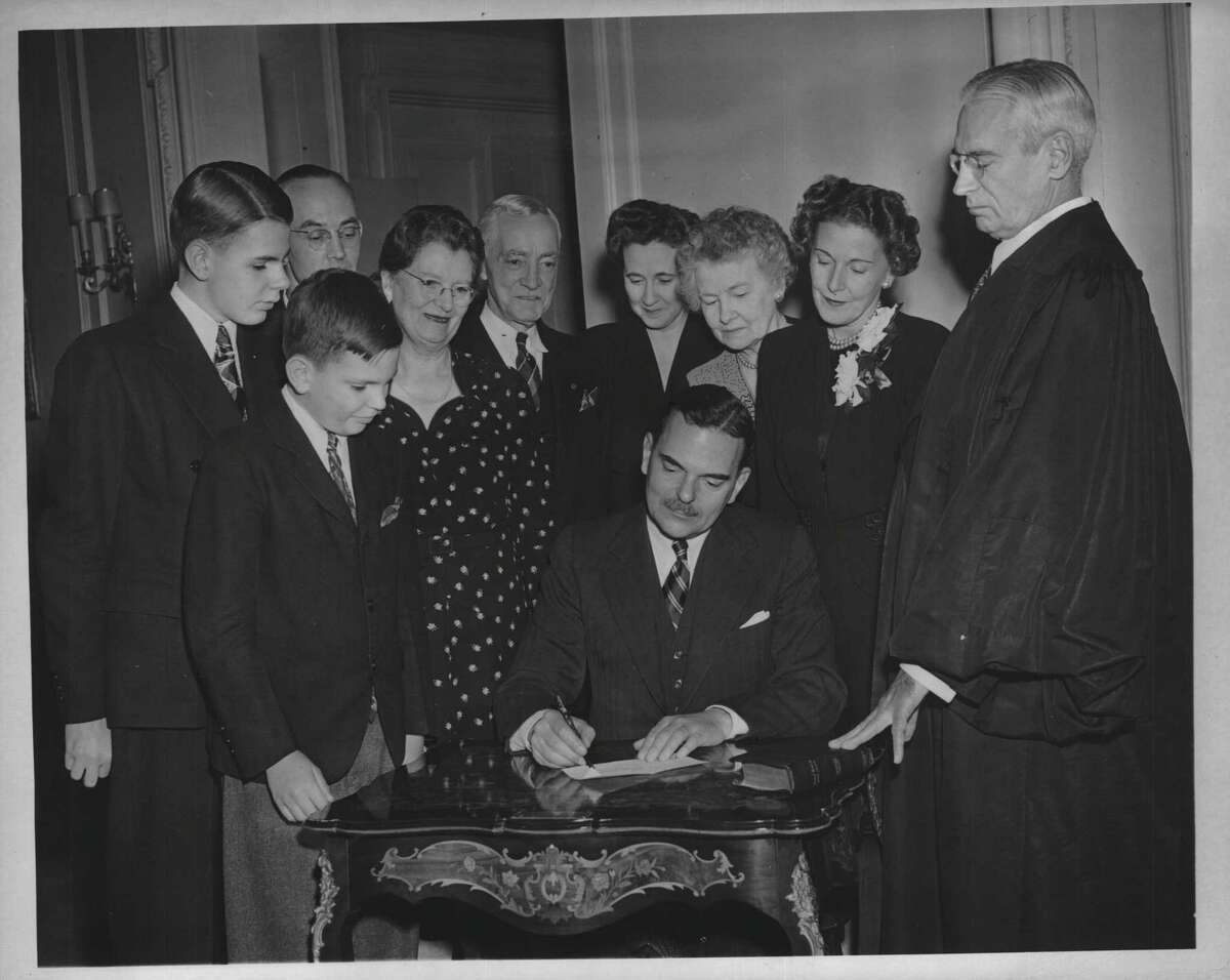 Gov. Thomas Dewey signs the official register, making him governor of New York State for a second term, at the Executive Mansion on New Year's Eve, Dec. 31, 1945. Looking on are John Dewey; Charles D. Hutt of Holly, Michigan, Mrs. Dewey's brother; Thomas Dewey Jr.; Mrs. George M. Dewey of Owosso, Michigan, the governor's mother; C.T. Hutt of Sapulpa, Oklahoma, Mrs. Dewey's father; Mrs. Charles D. Hutt of Holly, Michigan; Mrs. O.T. Hutt; Mrs. Thomas Dewey; and Judge John T. Loughran.