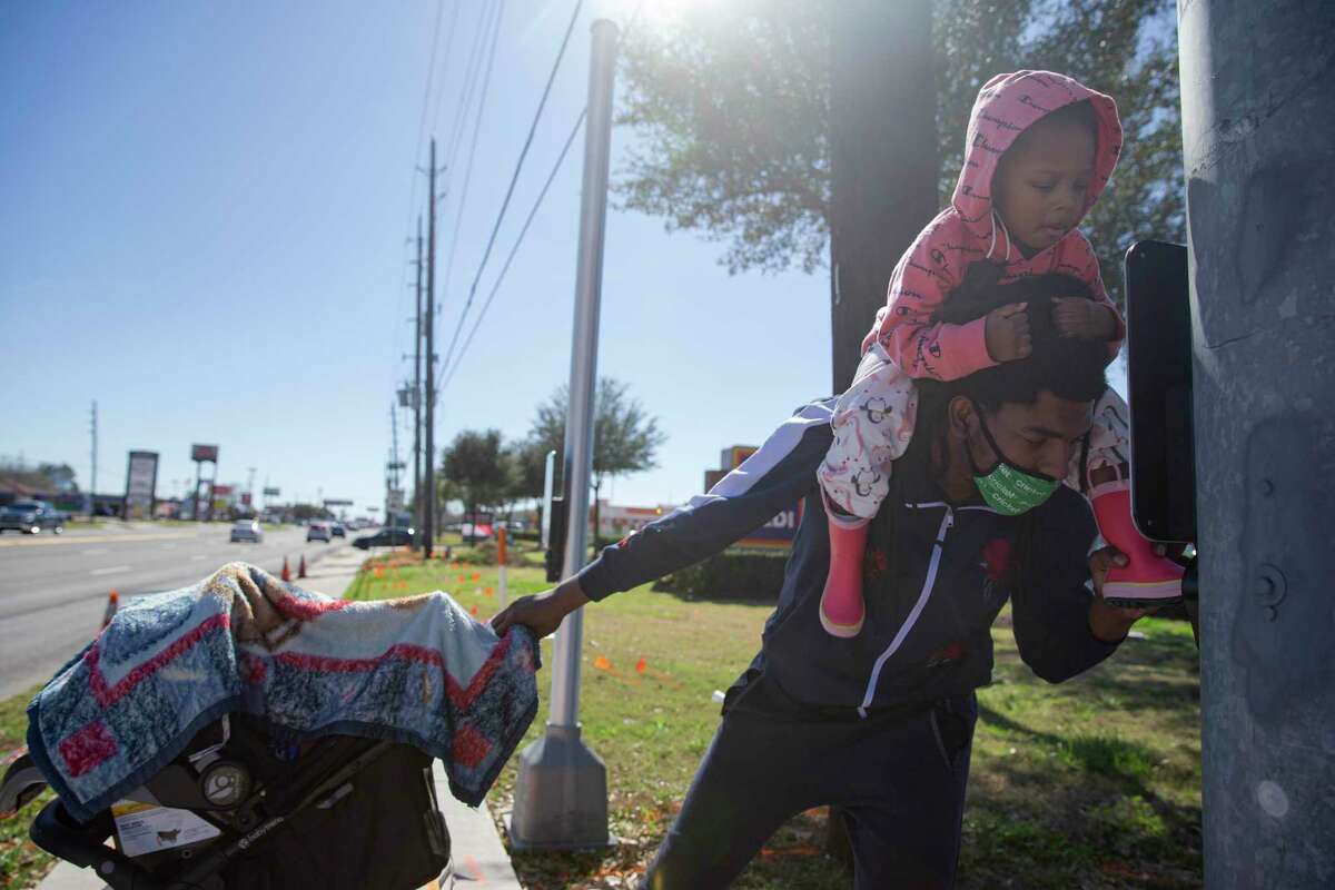 Derron Trent lets his older daughter, Naomi, 3, push the pedestrian crossing button to cross Texas 6 at Yorktown Crossing Parkway on Feb. 1, 2021, in Houston. Trent has been living in this area for almost two years, and before the sidewalks at the intersection were installed he said he had to push Naomi on the road.