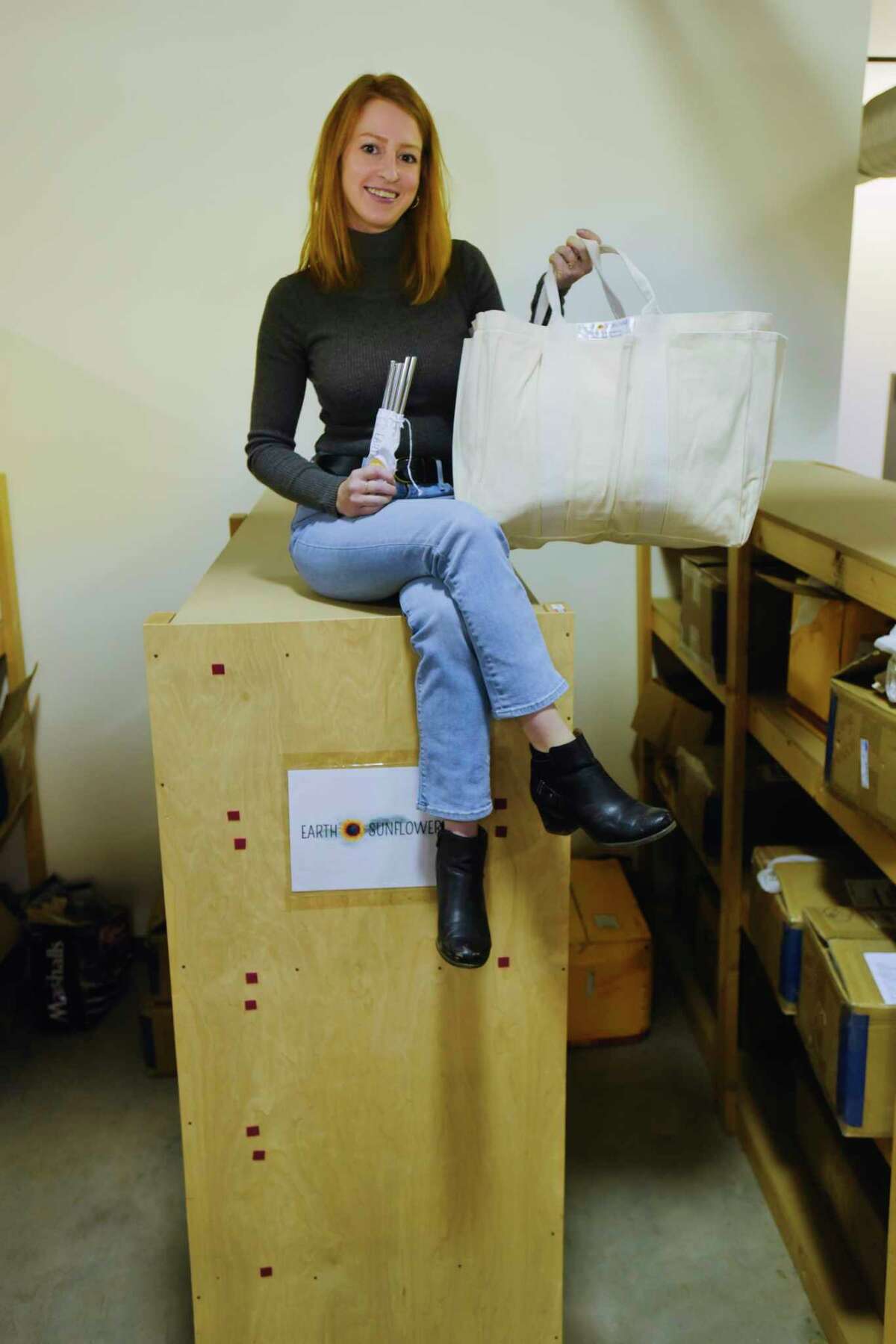 Jill Fecteau, owner of Earth Sunflower, sits on top of shelves holding boxes of the products she sells inside a warehouse where she rents space on Wednesday, Jan. 27, 2021, in Saratoga Springs, N.Y. Fecteau is holding stainless steel straws, which she started her business with, and a certified organic cotton extra large grocery bag with outside pockets, one of her bigger selling items currently. (Paul Buckowski/Times Union)