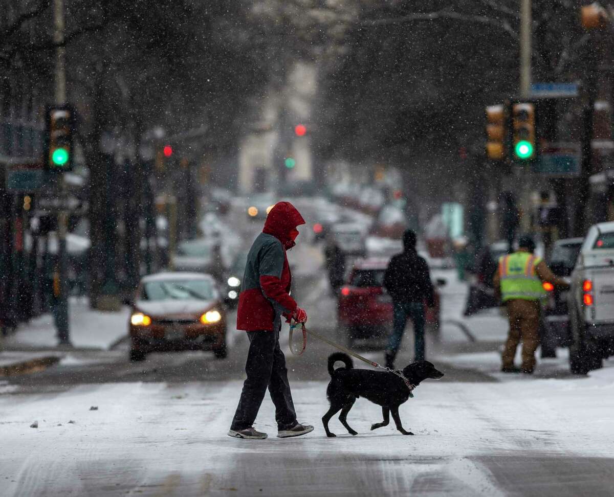 A person and their dog cross Houston Street Thursday, Feb. 18, 2021 as snow grips the city for the second time in a week, the result of an arctic air mass that sent temperatures plummeting and resulted in rolling blackouts across the city and the state. At one point about 1/3 of all CPS Energy customers were without electricity.