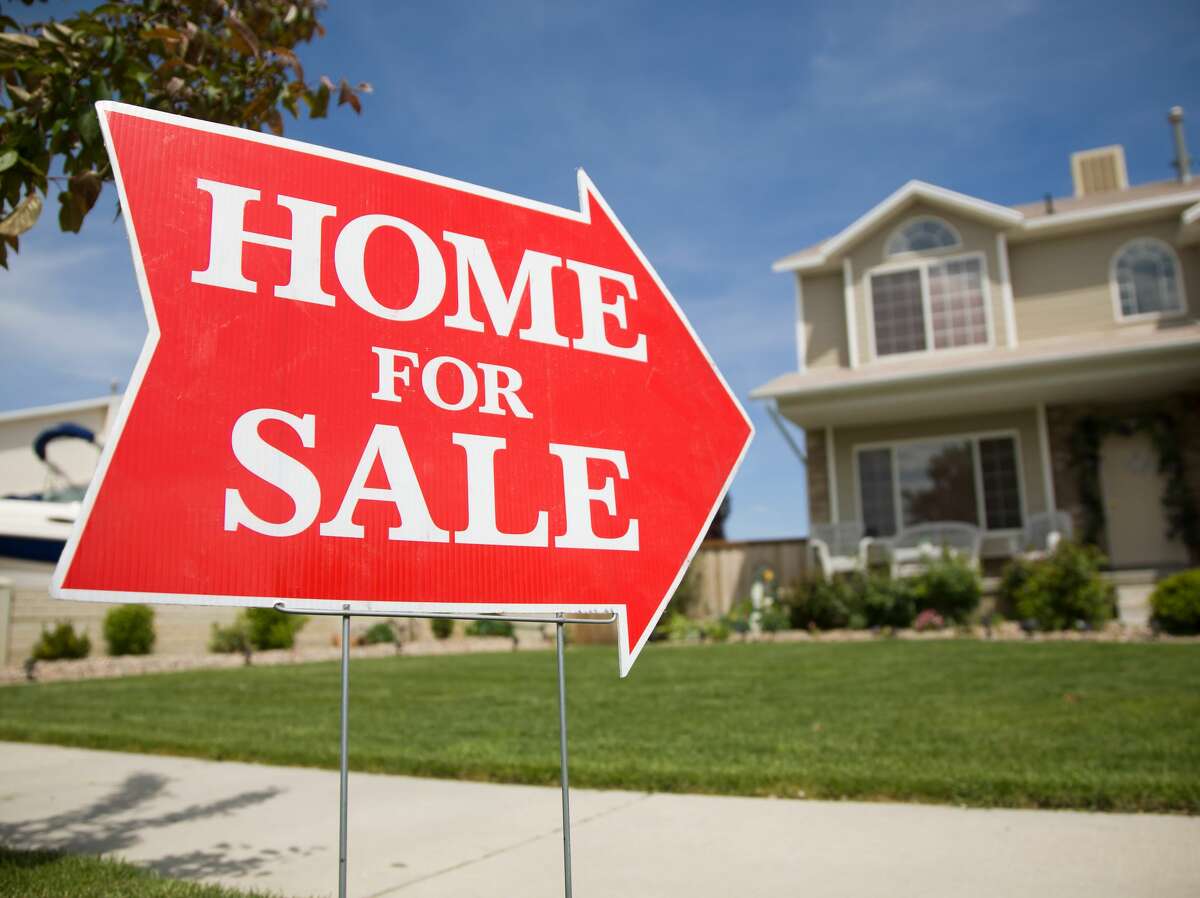 An arrow shaped red "Home For Sale" sign in front of a suburban 2-story home. The green grass and blue sky is visible in the background.