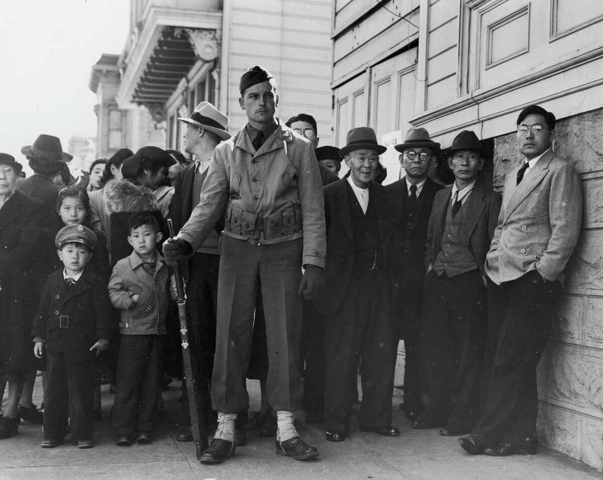 Forlorn looking American soldier standing guard in front of some Japanese American citizens awaiting transport to relocation camps after they were forcebly rounded up from their homes all along the west coast.