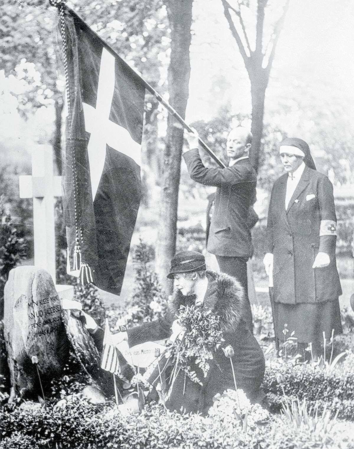 Kneeling before the tomb of a Danish war hero of 1895, Ruth Bryan Owen, first diplomatic minister of the United States, places a wreath and an American flag before the headstone of the patriotic shrine. The flag of Denmark flies overhead as a Danish Red Cross nurse (right) watches the ceremony.