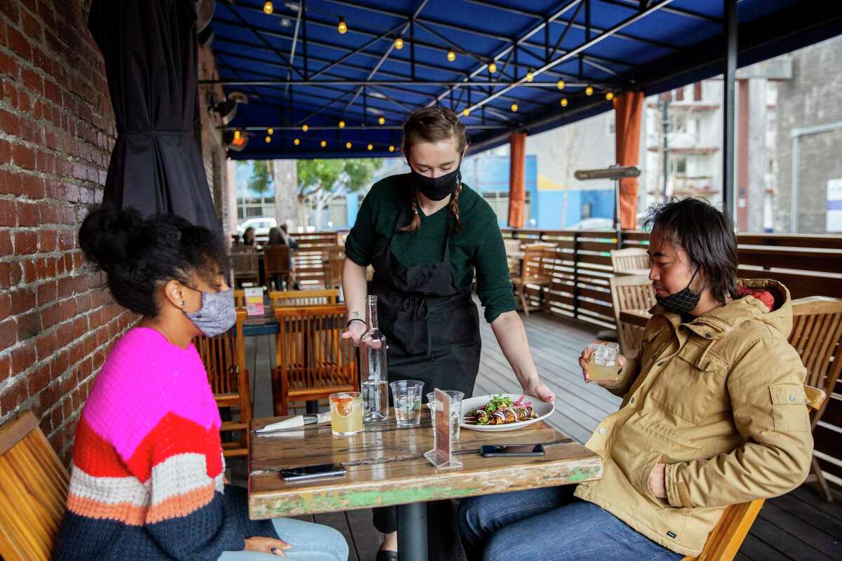 Maddie McGraw serves customers Rachel Kigano (left) and Ryohei Hinokuma at the Calavera patio in Oakland. Alameda County now has moved to the second-least restrictive tier of California’s reopening plan, allowing expansion of what activities can occur.