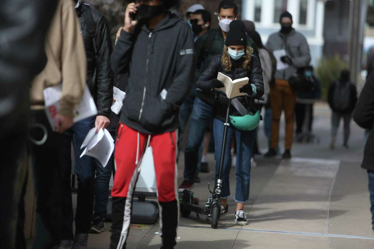 Mathilde Pons reads a book as she holds her scooter as she waits with her husband, Pierre Chesnot (behind Pons) in the line for drop-ins at the vaccine clinic at Zuckerberg General Hospital to recievie the COVID-19 vaccine on April 13, 2021 in San Francisco, Calif.