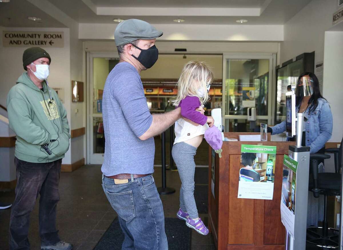 2021年4月20日星期二，加利福尼亚州奥克兰，保罗·霍特林(从左至右)在检查站抱着2岁的女儿西拉·霍特林(Sierra Houghtaling)拿着免提温度计。
