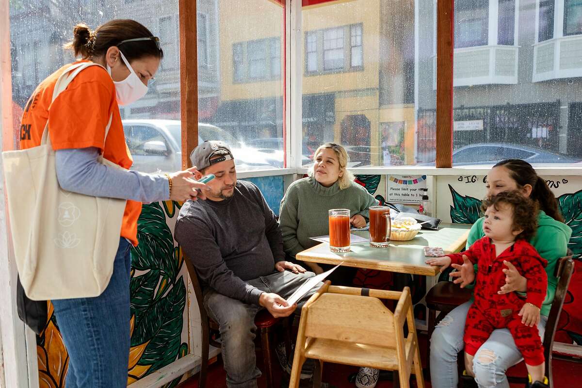 Michelle Tapia (center), Fernando Barajas, and their children have lunch while Latino Task Force and United in Health volunteer Noelle Bermingham uses her phone to register Michelle Tapia for a COVID-19 vaccine appointment on Saturday, April 10, 2021, in San Francisco, Calif.