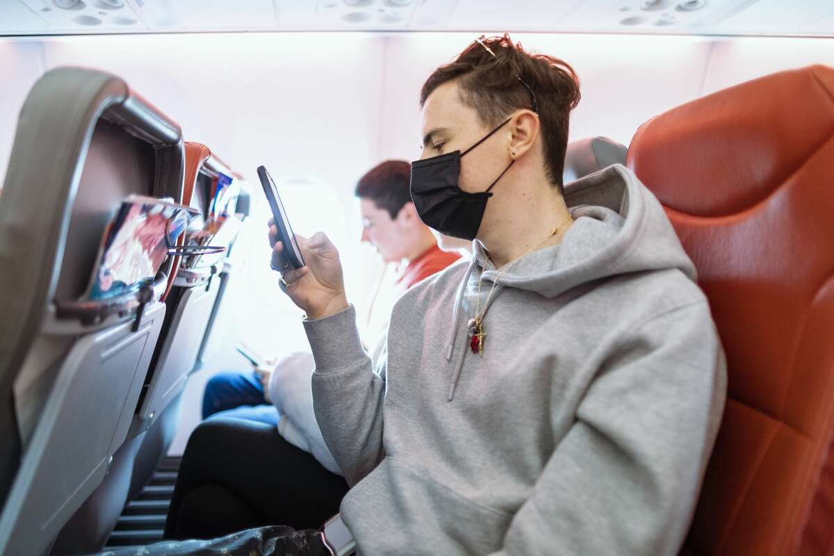 Man sitting on an airplane using his phone while wearing a surgical mask.