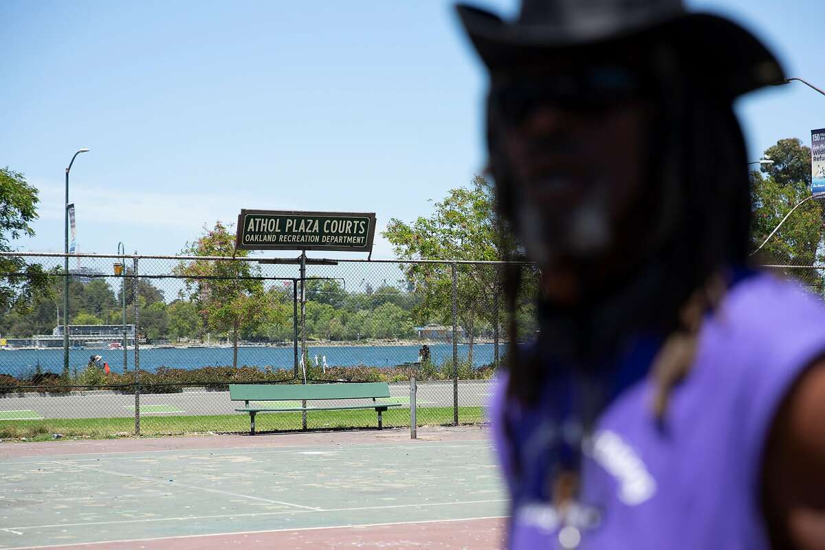 A sign reading Athol Tennis Courts is seen behind Nino Parker as he stands next to his tent and bike repair space in Oakland. He has been spared the crackdown on encampments in the city so far.