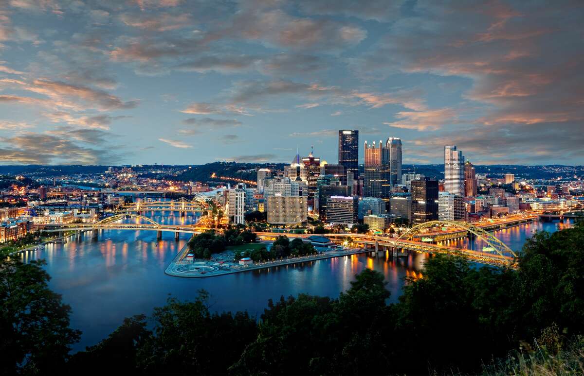 Horizonte de Pittsburgh, Pensilvania, EE.UU., al anochecer, con la confluencia de los ríos Monongahela y Allegheny en Point State Park formando el río Ohio en primer plano y edificios altos en el fondo.
