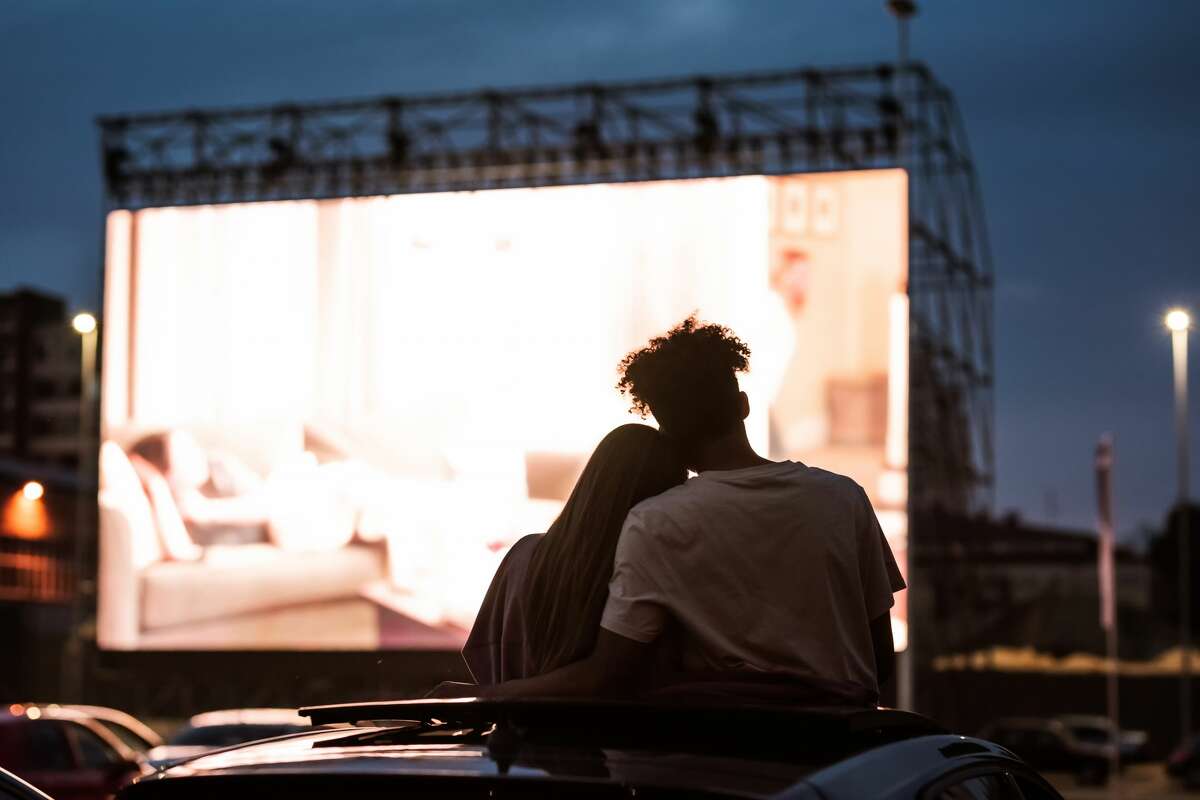 Vista ombreggiata di giovane coppia attraente, ragazzo e ragazza che si abbracciano, trascorrono del tempo insieme, seduti in macchina mentre si guarda un film in macchina al cinema.  Intrattenimento, concetto di incontri.  vista posteriore