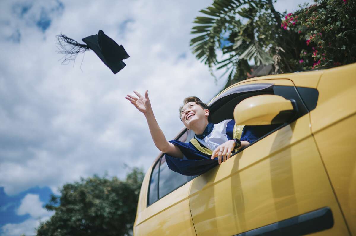 Young female with graduation robe throwing cap in the air from driver seat of her car laughing