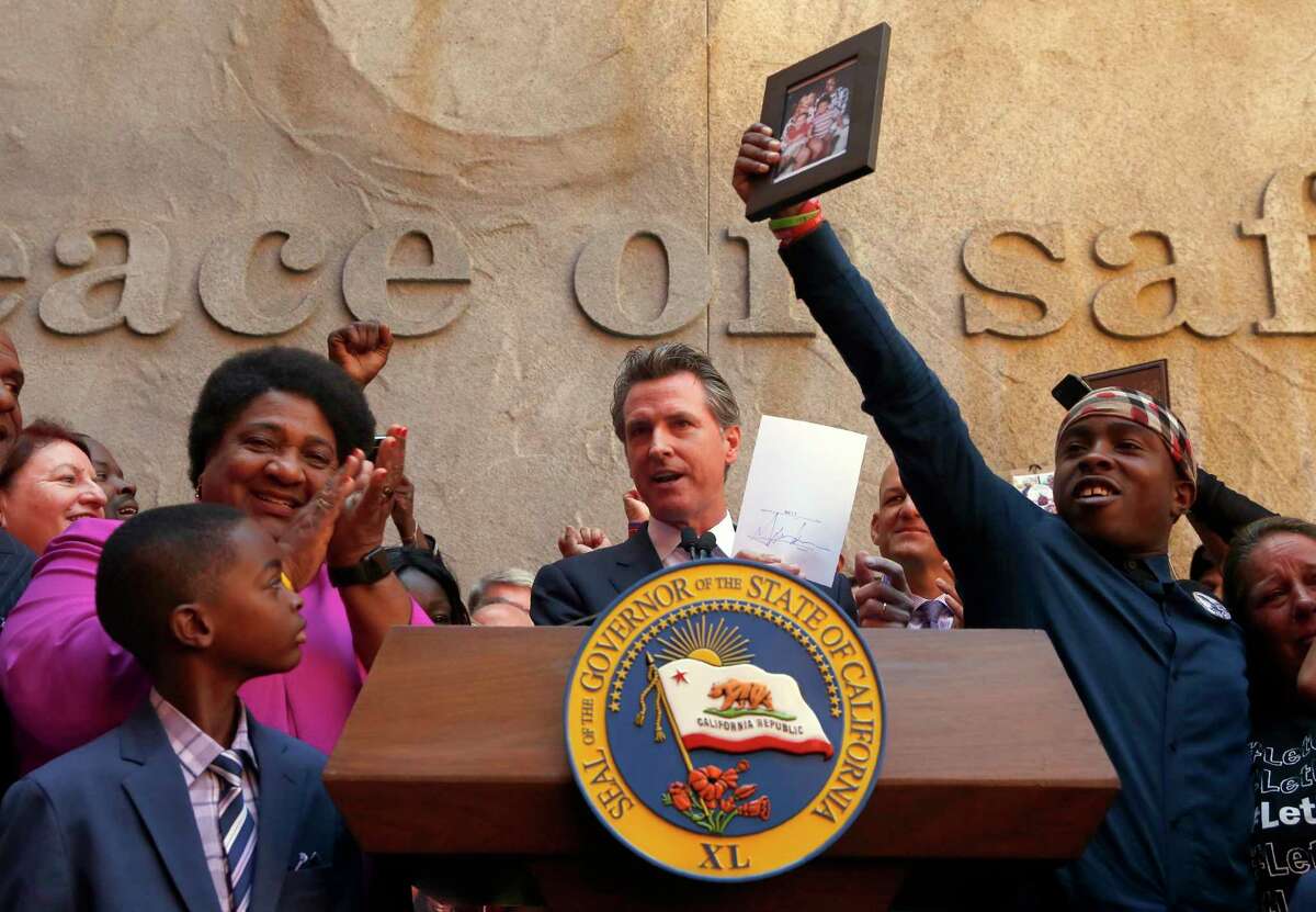 Gov. Gavin Newsom holds up the measure to limit police officers’ use of force by Assembly Member Shirley Weber, D-San Diego (left) after he signed it in August 2019, while Stevante Clark (right) holds up a photo of his brother, Stephon Clark, who was shot and killed by Sacramento Police in 2018.