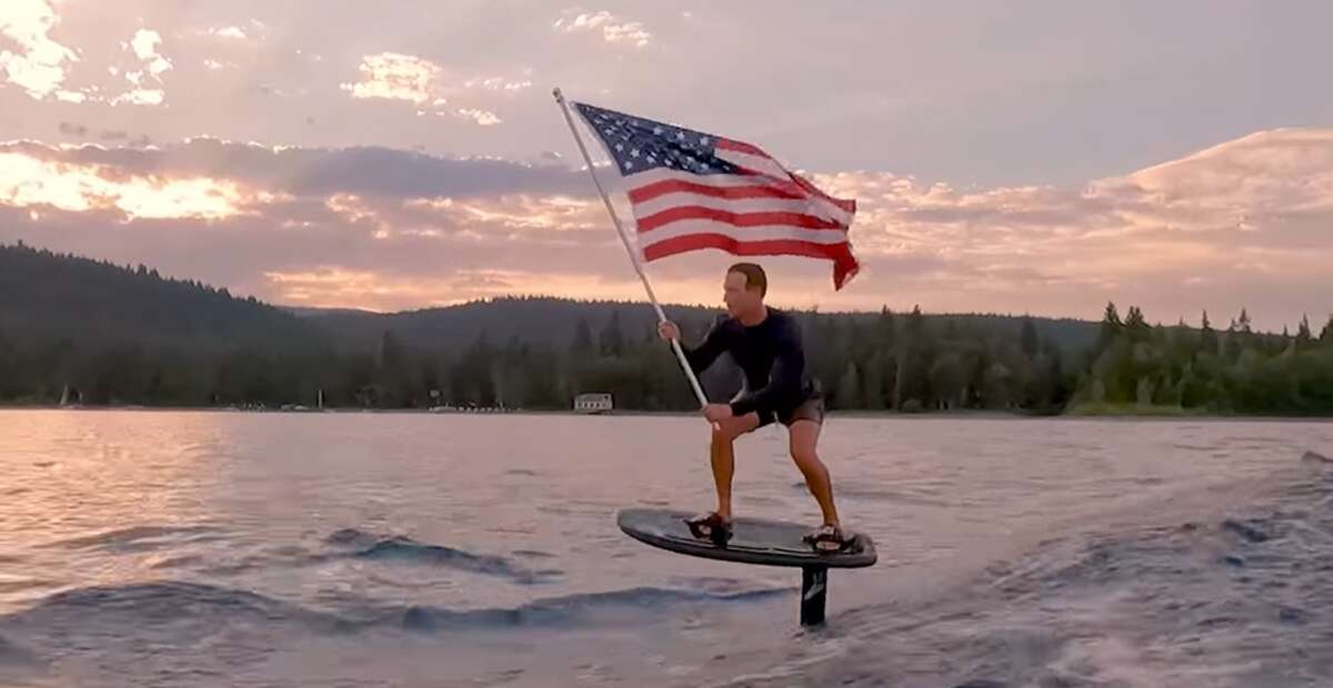 Mark Zuckerberg rides an electric hydrofoil surfboard waving a US flag.