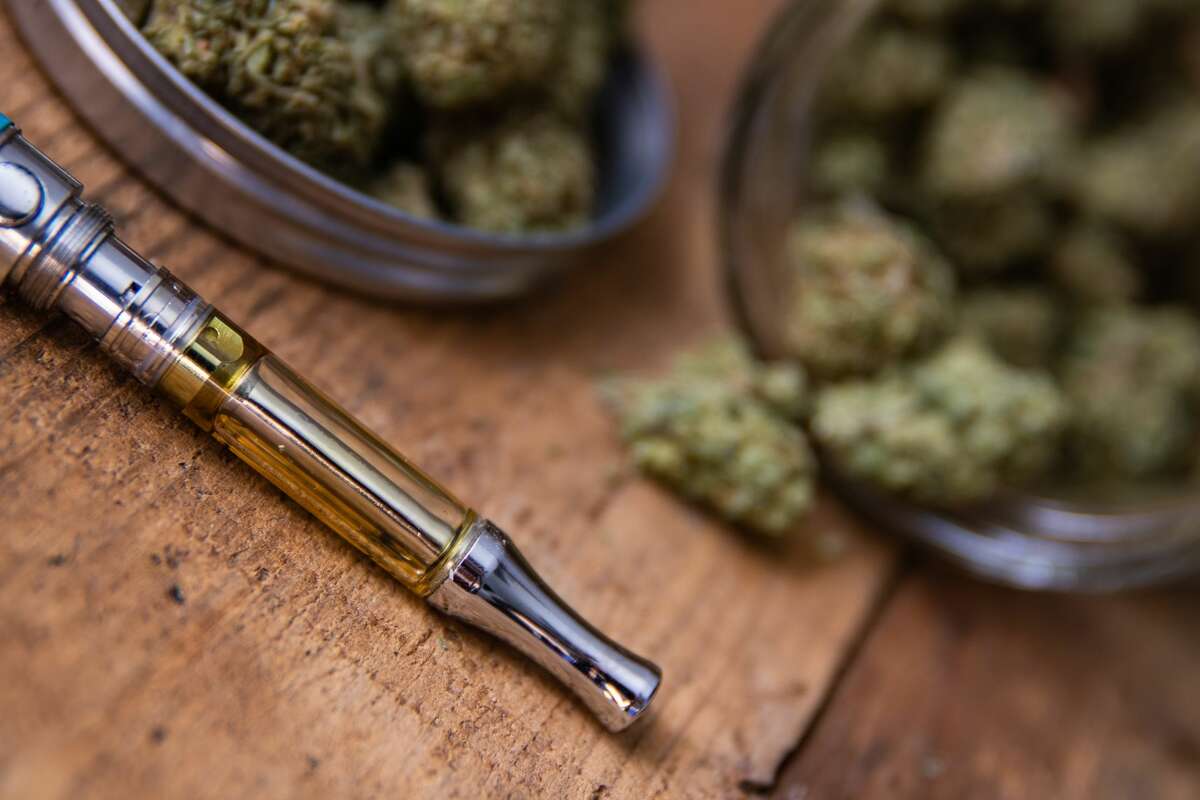 THC oil concentrate filled vape pen on natural wood with an open glass container full of marijuana buds grown and sold in dispensaries throughout the state.