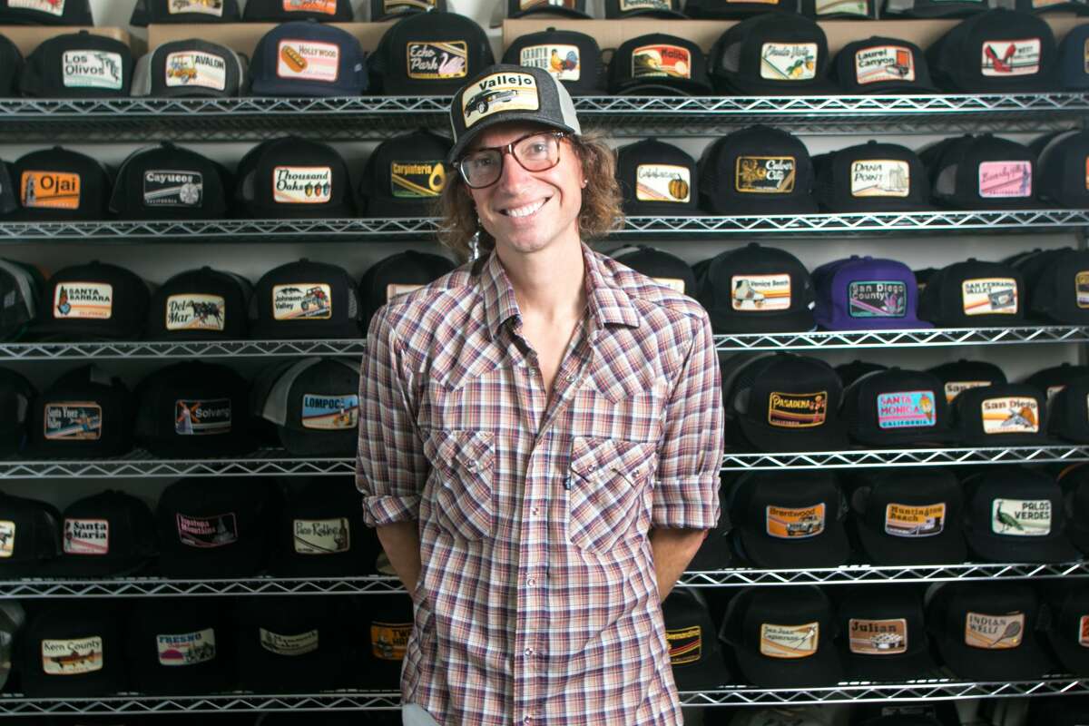 Owner and lead designer Luke Fraser poses in front of hats adorned with patches his company, Bart Bridge, designed at their office and warehouse located inside a house in Vallejo, Calif., on Aug. 17, 2021. The company makes patches that celebrate small towns, particularly in the San Francisco Bay Area and California, then puts them on hats for sale.