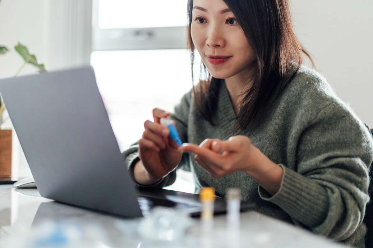 Young woman using a blood test kit at home while doing health check and consultation online. Home finger-prick blood test.
