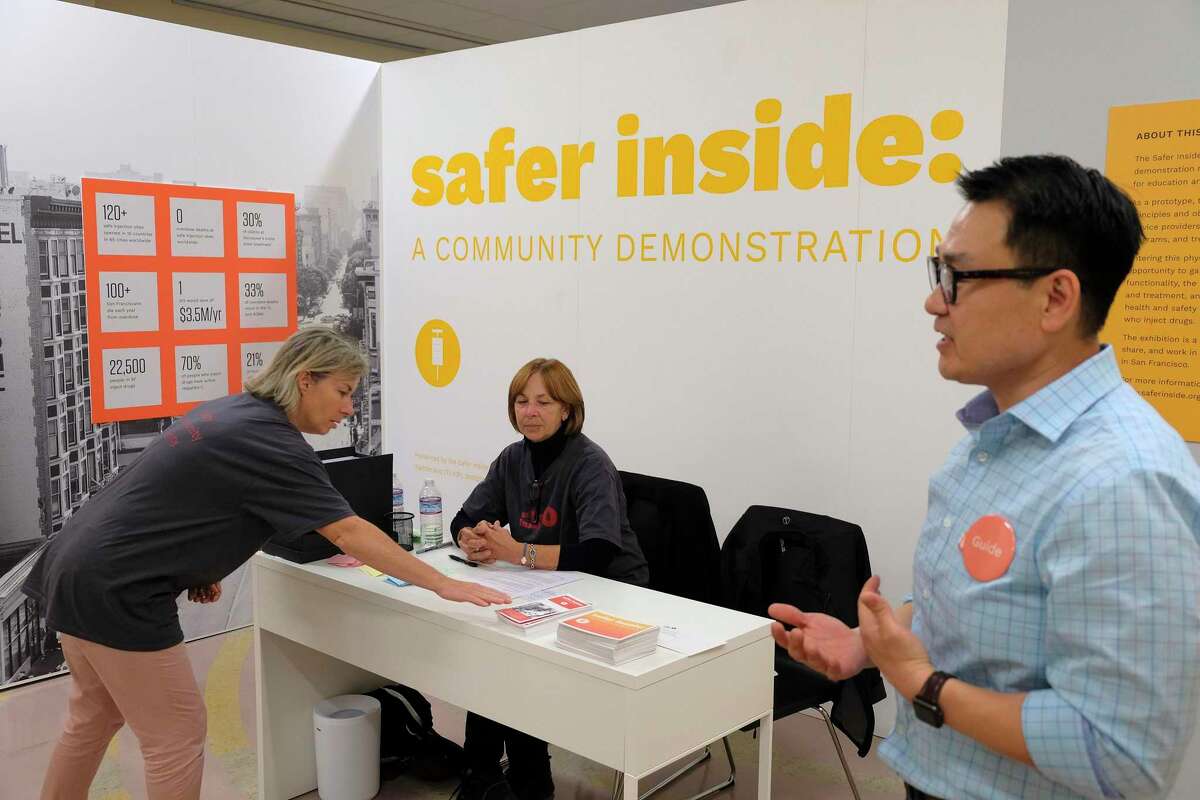 In this photo taken Wednesday, Aug. 29, 2018, Marie Fauvel, left, and Pat Shea, center, talk at a reception desk at Safer Inside, a realistic model of a safe injection site in San Francisco. At right giving a tour is clinical director Kenneth Kim. The model is an example of a supervised, indoor location where intravenous drug users can consume drugs in safer conditions and access treatment and recovery services. (AP Photo/Eric Risberg)