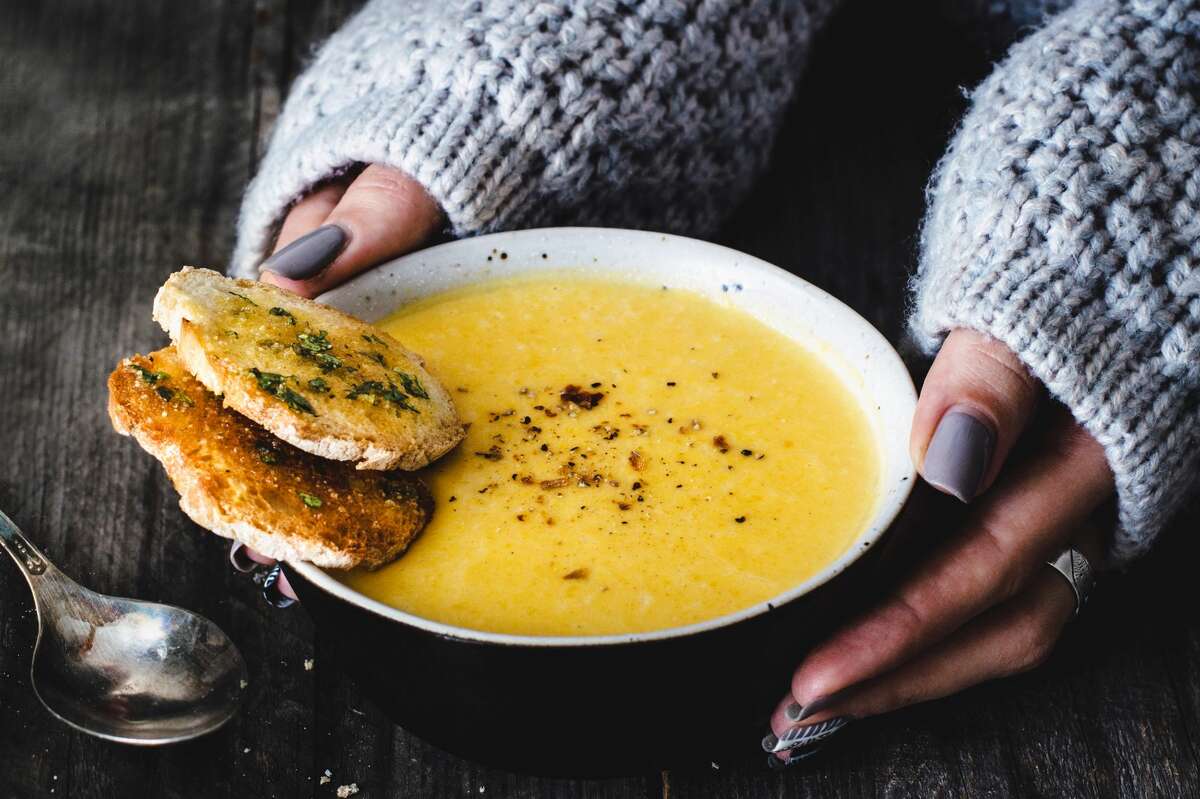 Woman hands holding bowl of warm carrot pumpkin cream soup served with garlic bread on rustic wooden table.