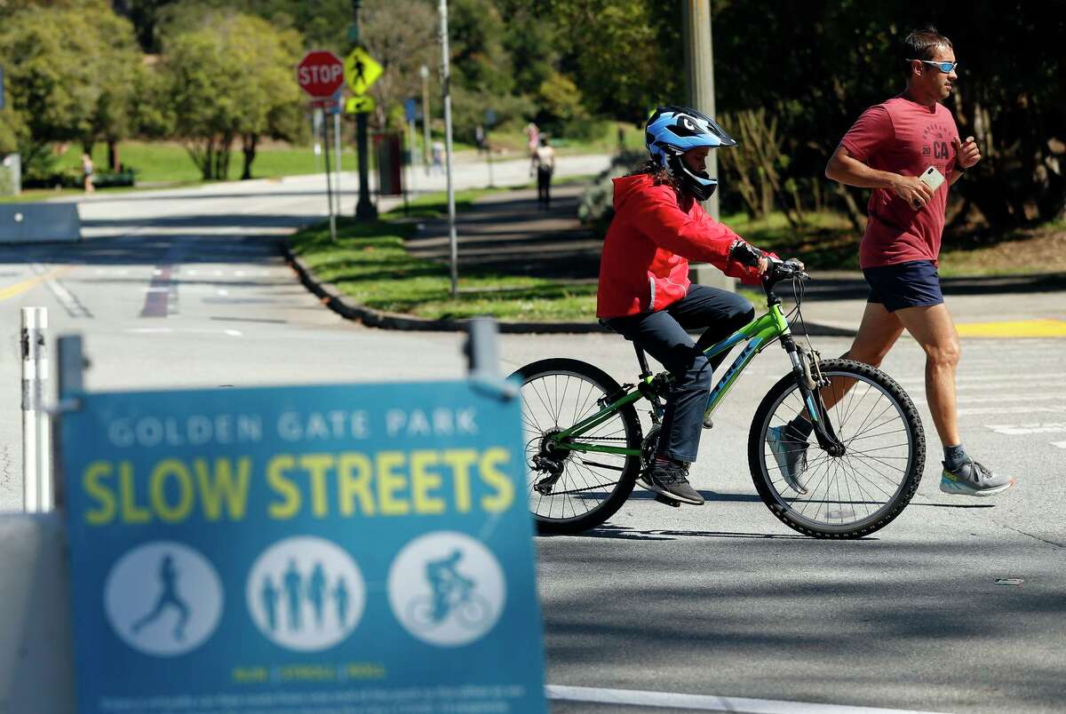 A jogger and biker pass a Slow Streets sign at John F. Kennedy and Hagiwara Tea Garden drives in Golden Gate Park. JFK Drive, which was closed to cars during the pandemic, is the center of a debate on whether to allow cars in the future.