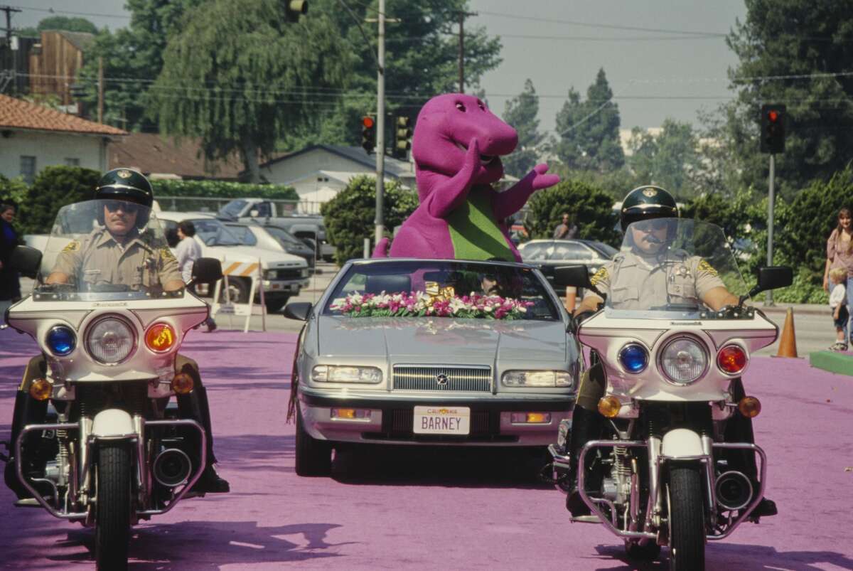 Barney, the purple Tyrannosaurus rex from children's television series 'Barney & Friends', riding in a convertible car following behind police outriders, at an unspecified event, location unspecified, circa 1995. (Photo by Vinnie Zuffante/Michael Ochs Archives/Getty Images)