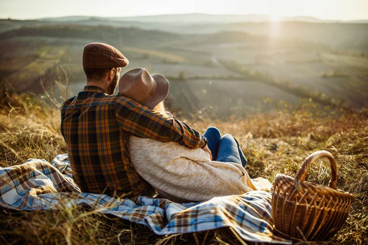 Back view of embraced couple relaxing on picnic in autumn day and looking at view.