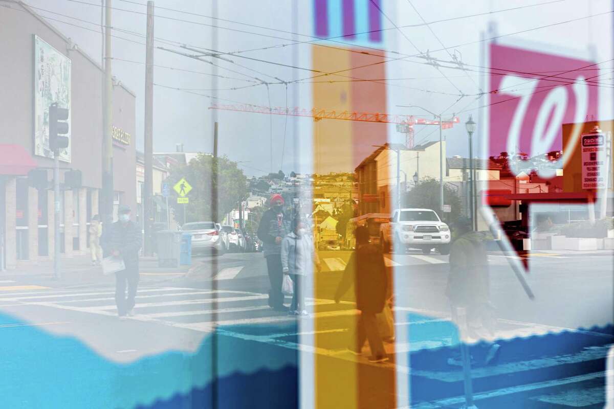 Pedestrians crossing the street are seen in a reflection on the window of the Walgreens at 4645 Mission St.