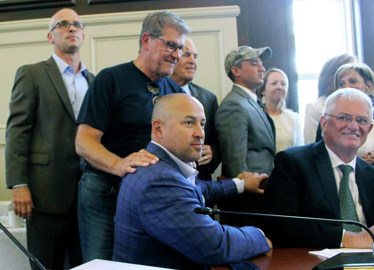 University of Connecticut men's basketball coach Dan Hurley, left, women's basketball coach Geno Auriemma, second from left, and athletic director David Benedict, seated foreground, watch the board of trustees vote on Wednesday, June 26, 2019, on the school's campus in Storrs, Conn., to move most of UConn's athletic teams from the American Athletic Conference to the Big East.