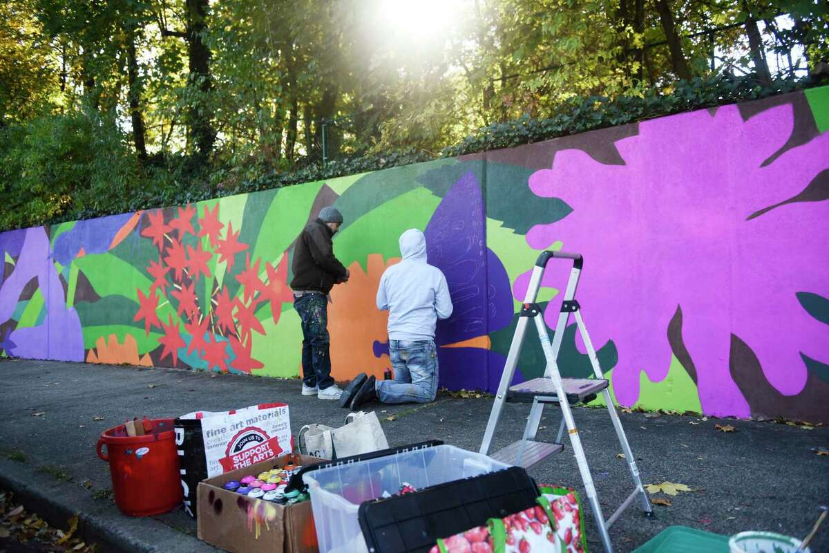 Artist Nelson Rivas of Newburgh, N.Y., left, and his assistant, Yedi, paint a mural on the wall behind the bus stop near Greenwich High School in Greenwich, Conn. Wednesday, Nov. 3, 2021. He was commissioned by the Greenwich Conservation Commission to paint the mural of colorful pollinator pathway flowers.