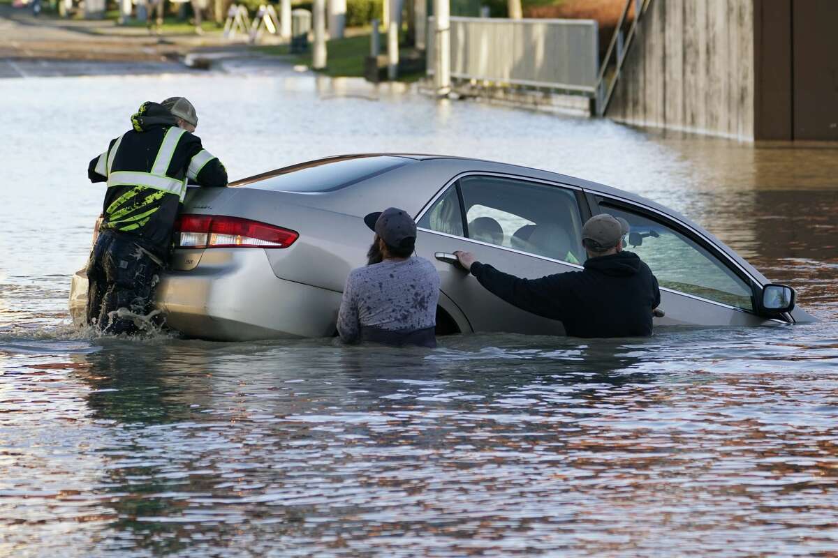 One man jumps on the back of car to try to keep the front from sinking further as others surround it, with the driver still aboard, that went into the flooded Nooksack River on Main Street, Tuesday, Nov. 16, 2021, in Ferndale, Wash. The group of citizens went into the river and stopped the car from floating further or sinking, whose driver had gone past a road closed sign, then muscled it back onshore. No one was injured. (AP Photo/Elaine Thompson)