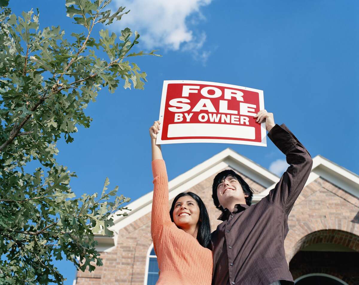 A couple holds a "For Sale By Owner" sign in front of a home. 