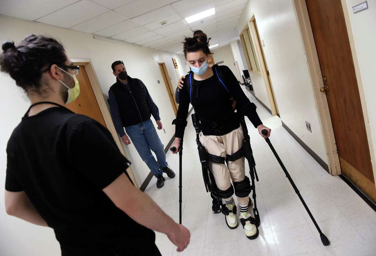 Nick Matarazzo, center, of Deep River watches his daughter, Natalie, 18, who suffered a spinal cord injury, walk in a hallway of Gaylord Specialty Healthcare in Wallingford on Dec. 3, 2021,wearing an Ekso robotic skeleton. At left is rehab aid Will Bermender and behind Natalie is physical therapist Ingrid Marschner.