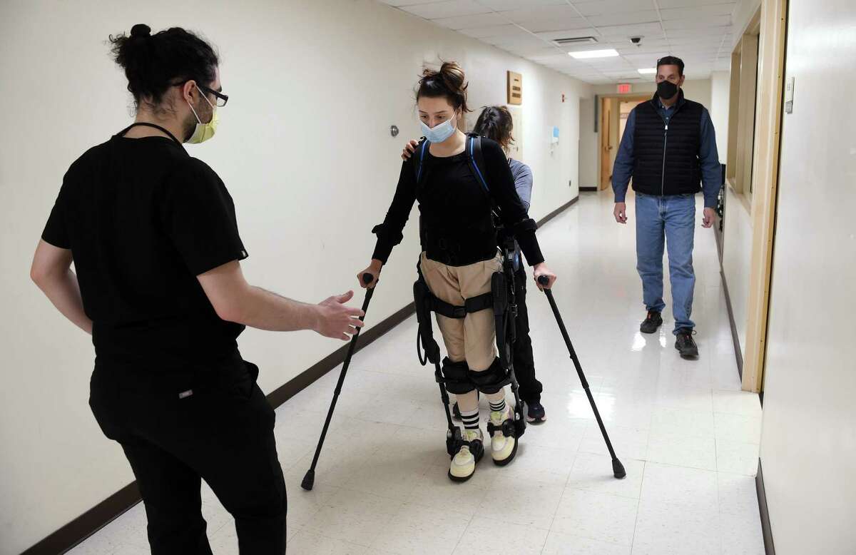 Nick Matarazzo, right, of Deep River watches his daughter, Natalie, 18, who suffered a spinal cord injury, walk in a hallway of Gaylord Specialty Healthcare in Wallingford on Dec. 3, 2021, wearing an Ekso robotic skeleton. At left is rehab aid Will Bermender and behind Natalie is physical therapist Ingrid Marschner.