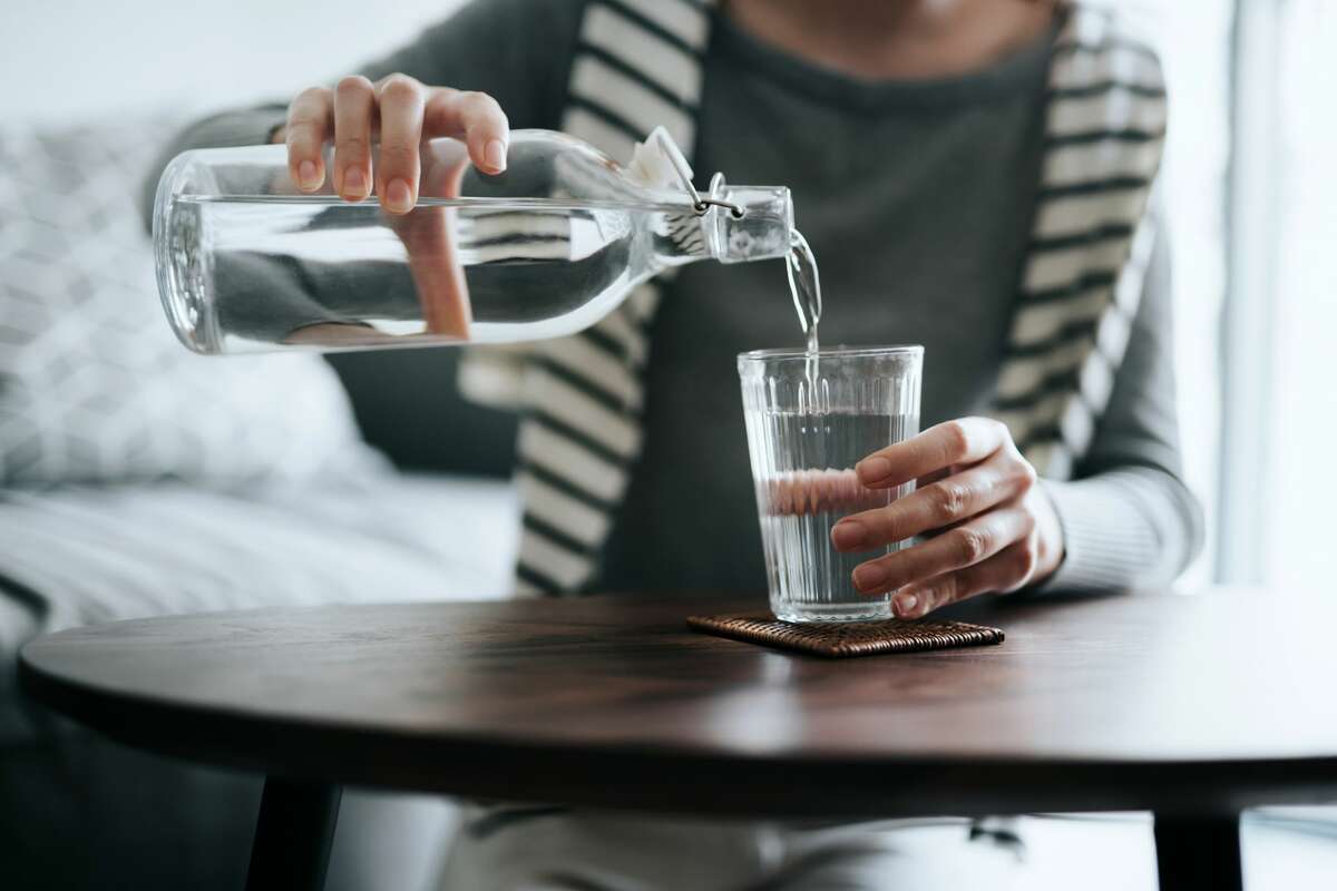 Close up of young woman pouring water from bottle into the glass on a coffee table at home. 