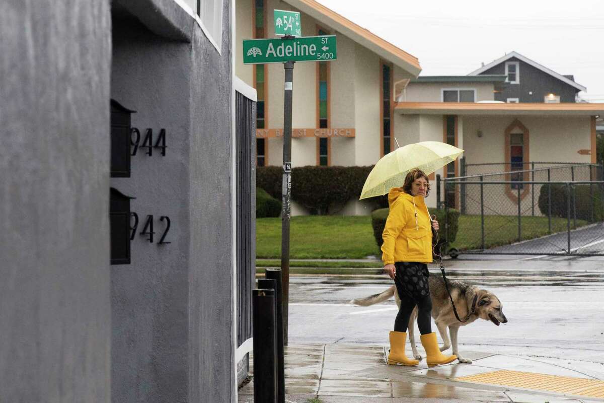 A woman wears a raincoat, boots and umbrella while walking her dog along Adeline Street in Oakland, Calif. as heavy rain falls across the region.