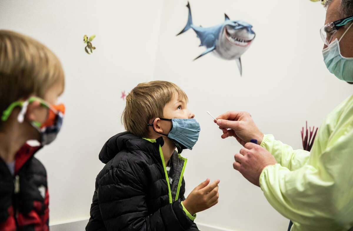 Joint caption: Above: Six-year-old Jimmy Brady, center, receives a COVID-19 PCR test from Dr. Joshua Parker, right, as brother Tucker, 4, looks on at Golden Gate Pediatrics in San Francisco, Calif. Tuesday, Dec. 28, 2021. Below: A tray of Pfizer COVID-19 vaccine is seen next to a tray of lollipops in an examination room used for COVID-19 vaccination at Golden Gate Pediatrics in San Francisco, Calif. Tuesday, Dec. 28, 2021.